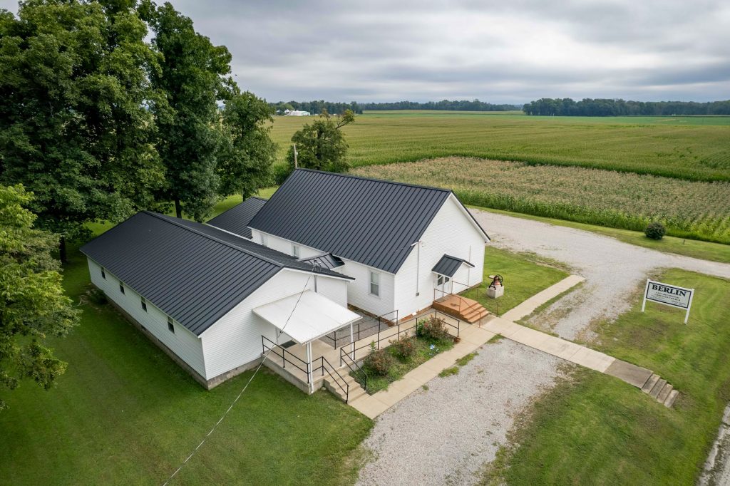 Aerial view of a white church featuring a standing seam black roof, surrounded by greenery and open space.