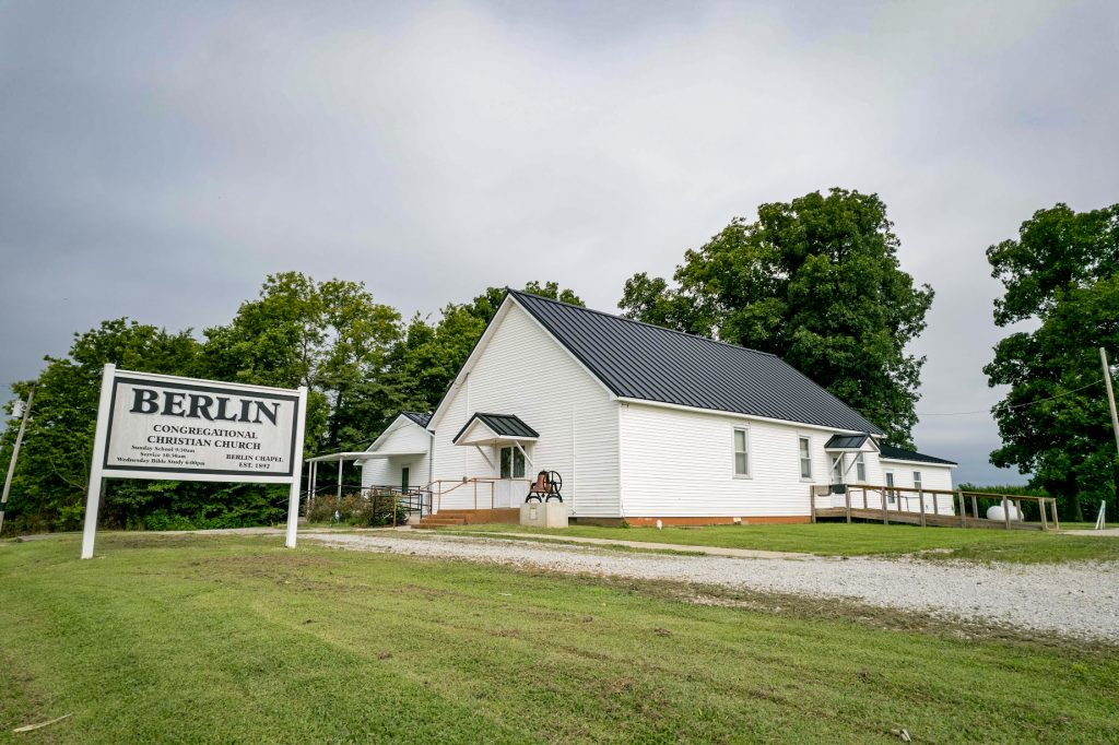 The Berlin Congregational Christian Church sign stands in front of their white church building with a standing seam roof.