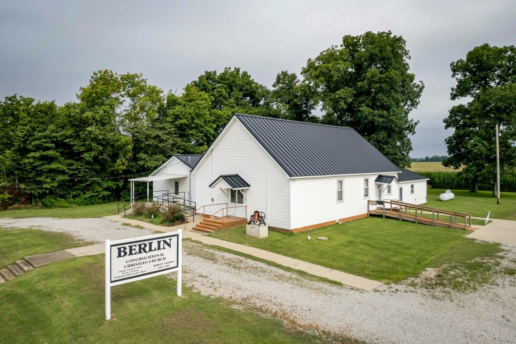 A church in a rural setting featuring a standing seam roof, nestled among greenery and farmland.