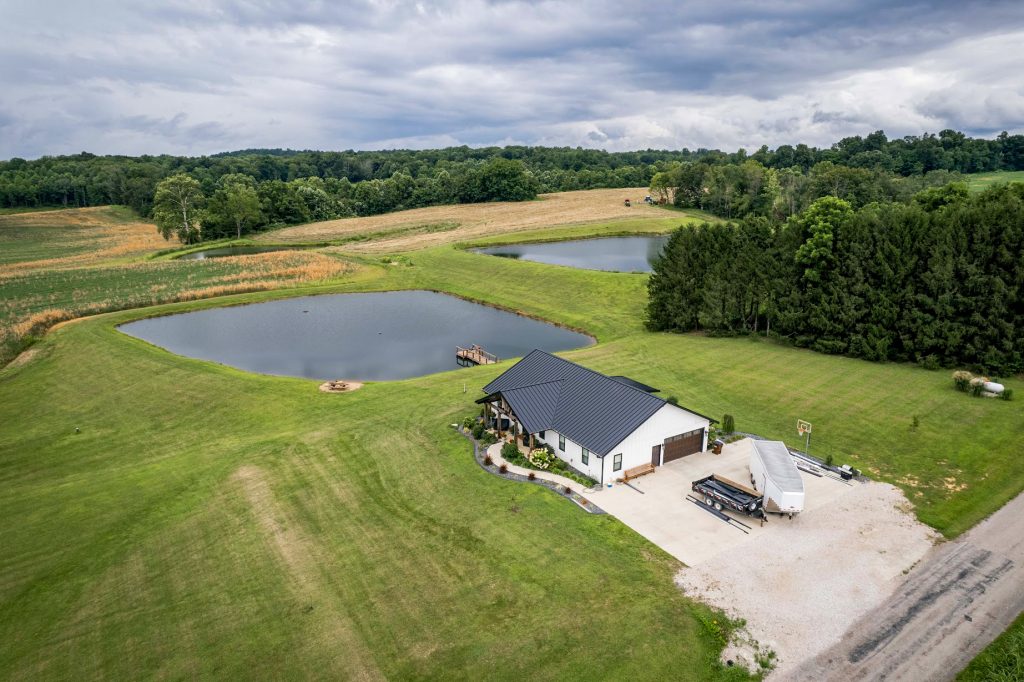 A serene setting that includes a calm pond and a house with a standing seam roof, situated in expansive fields.