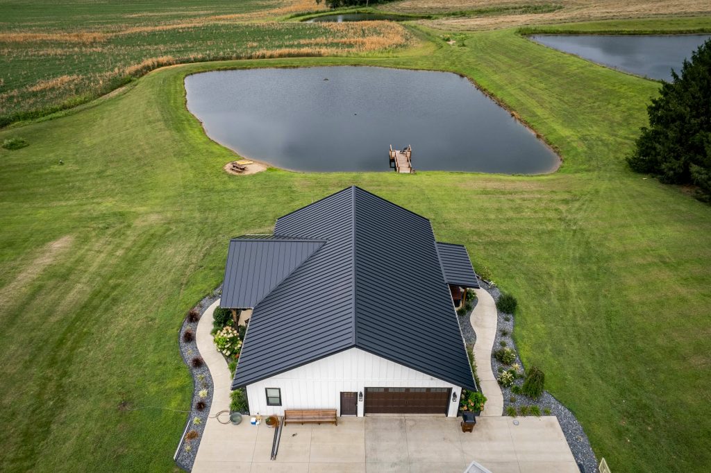 Aerial perspective of a house featuring a standing seam roof, adjacent to a tranquil pond.