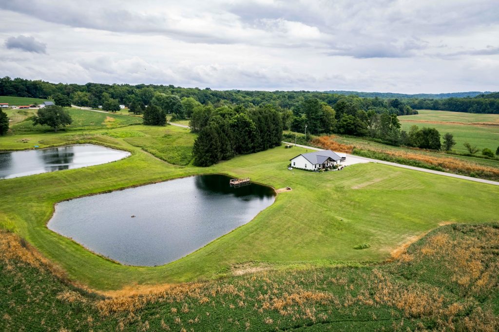 Aerial view of a farm featuring a house with a standing seam roof and a pond surrounded by fields.