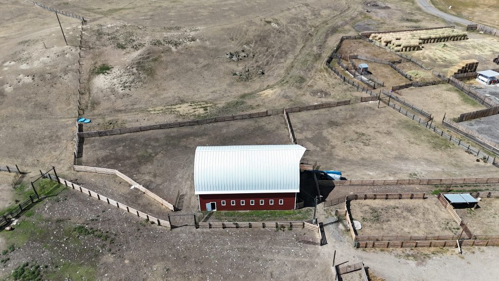 Aerial view of red horse barn with white curved arched metal roof surrounded by livestock corrals
