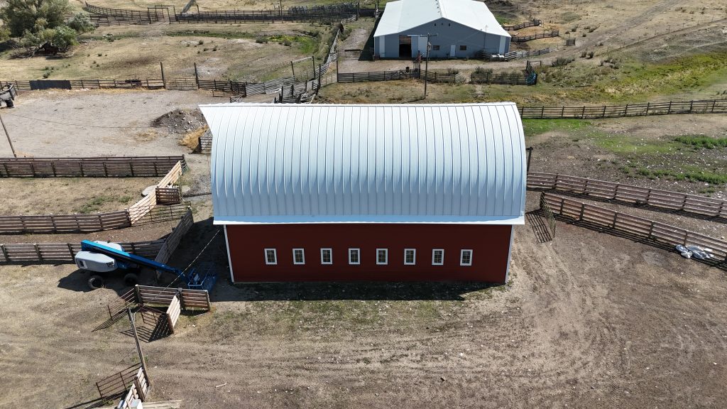 Red barn with white curved arched metal roof in working ranch setting with livestock corrals