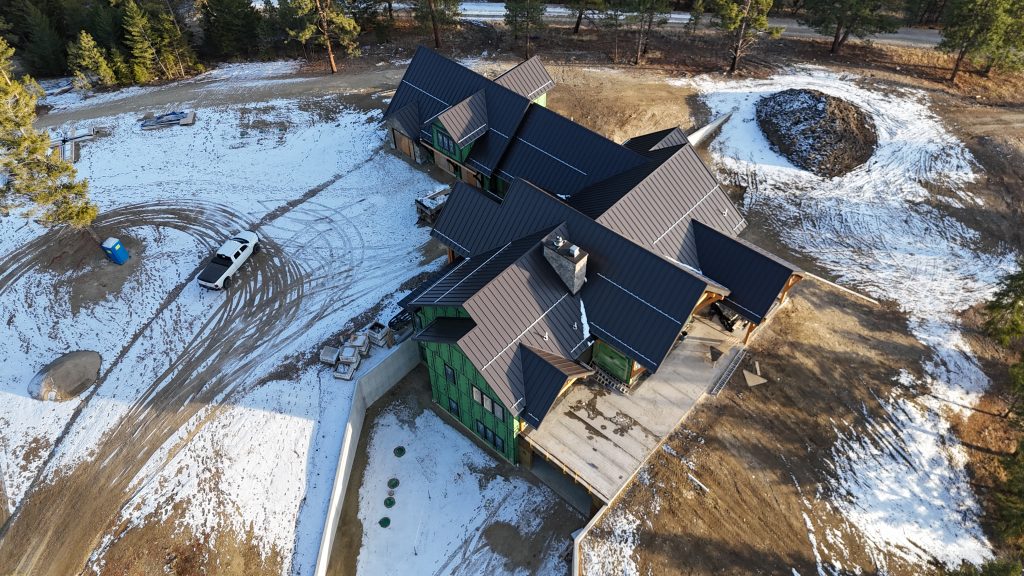 Winter aerial of custom home construction with completed dark metal roof in mountain forest setting