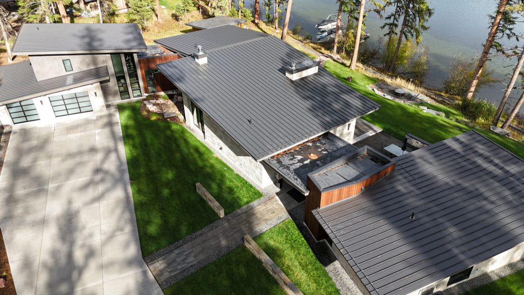 Angled aerial of modern lakefront home featuring charcoal metal roof and concrete driveway approach