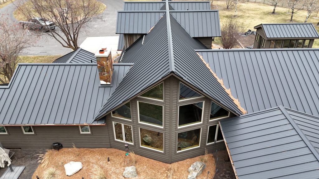 Close aerial of charcoal metal roof on ranch home highlighting large glass gable windows and stone chimney