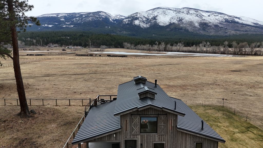 Barndominium with charcoal metal roof against panoramic snow-capped mountain backdrop and ranch fields