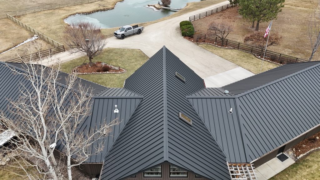 Angled aerial of ranch home with charcoal metal roof showing pond and circular driveway on rural property