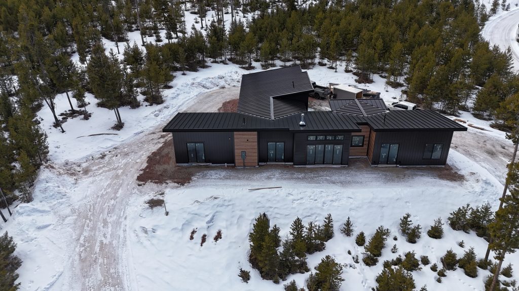 Aerial drone view of modern new construction home with matte black standing seam metal roof black board and batten siding cedar horizontal accent panels and large black-framed windows in snow-covered forested mountain setting