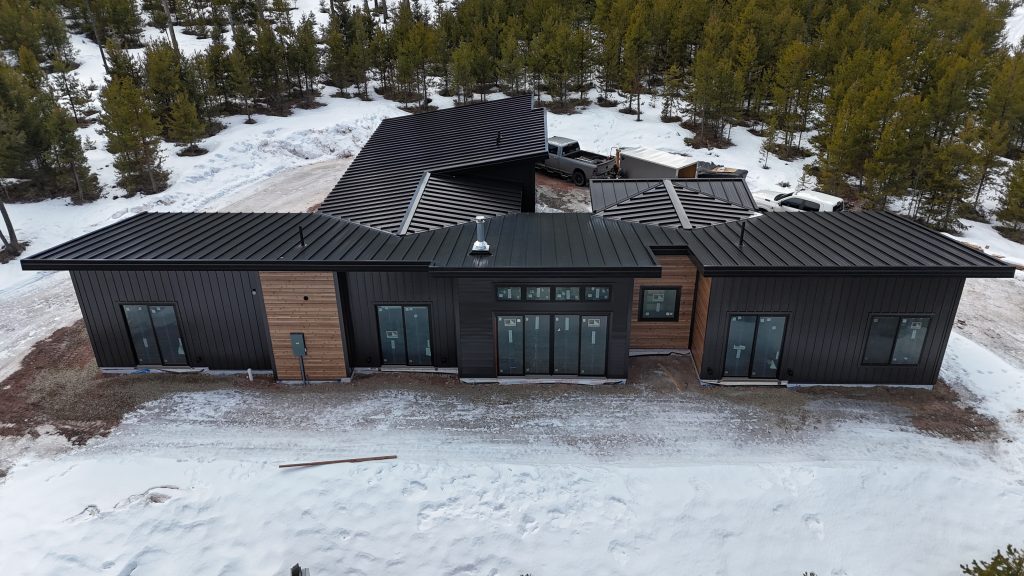 Aerial drone view showing complex multi-plane matte black standing seam metal roof geometry on modern home with black board and batten siding cedar horizontal accents and large glass door and window walls in winter