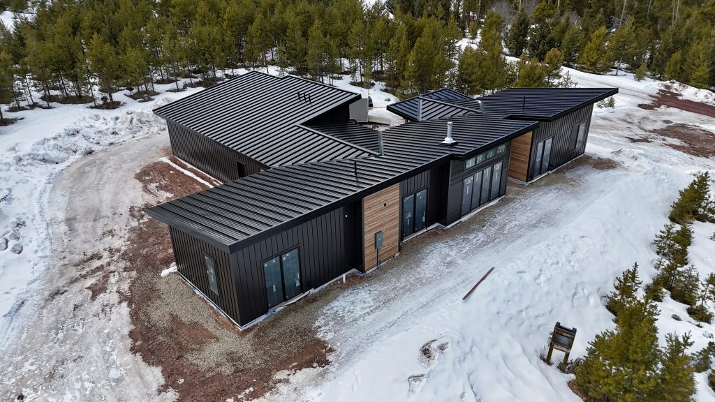 Aerial drone view of modern home under construction with matte black standing seam roof timber post entry canopy black board and batten siding cedar horizontal accent panels and crew trucks on site in winter