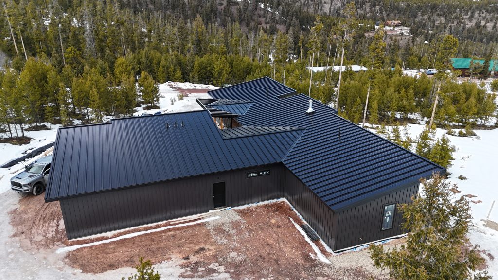 Aerial drone view of rear and side elevation of modern home with matte black standing seam multi-slope roof black board and batten siding and small window accents surrounded by snow-covered pine forest