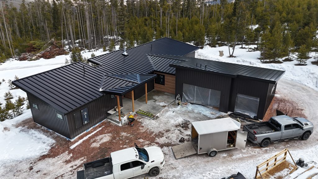 Aerial drone front elevation view of modern new construction home with matte black standing seam roof multi-plane rooflines black board and batten siding cedar horizontal accent panels and floor-to-ceiling glass door wall in winter mountain setting