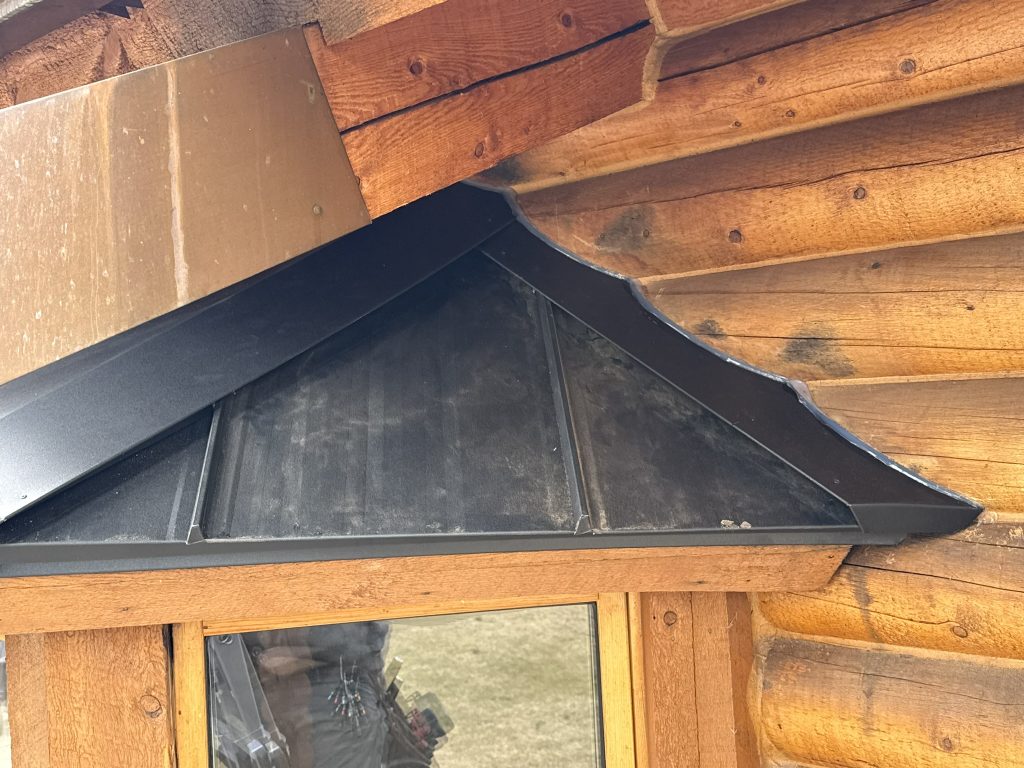Close-up detail of custom black metal roof cricket and flashing work above window on log cabin