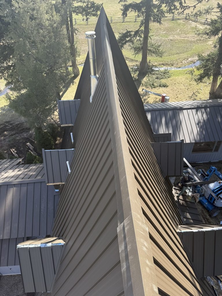 Ground-level view along standing seam metal roof ridge showing panel seams and stovepipe penetration during installation