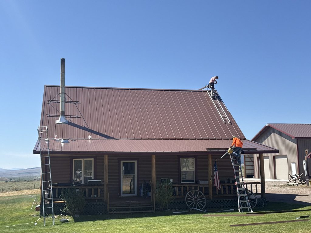 Two-man crew installing mahogany brown standing seam metal roof panels on rural log cabin home with clear blue sky and open ranch land in background