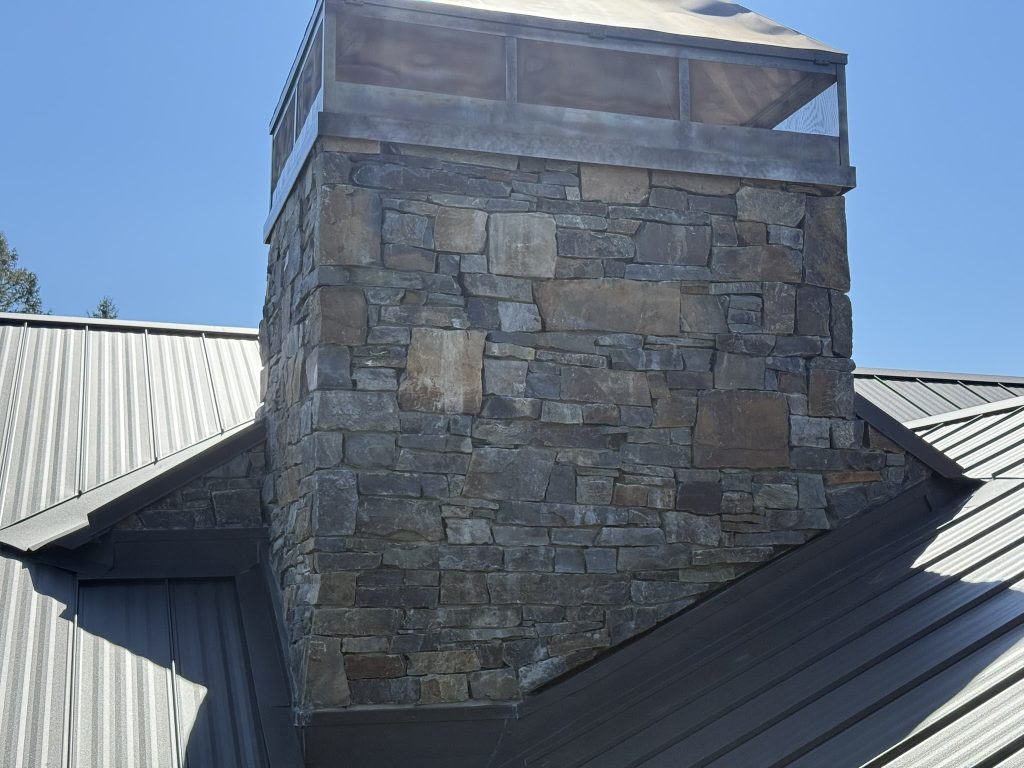 Close-up of large natural stone chimney with custom fabricated metal cricket and cap flashing on a dark gray standing seam metal roof