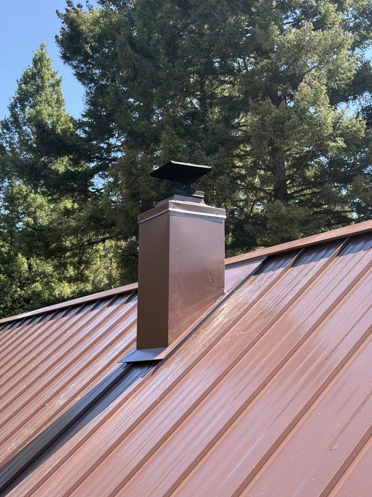 Completed metal chimney enclosure box finished to match copper brown standing seam metal roof panels with black spark arrestor chimney cap