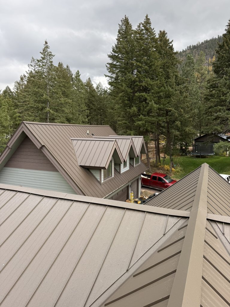 Elevated view of standing seam metal roof with integrated dormer windows and matching metal roof panels in tan and brown tones against forested mountain backdrop