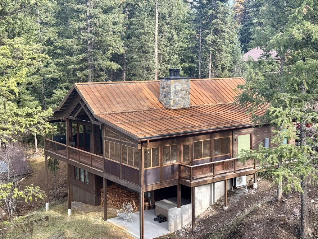 Aerial drone view of completed copper-finish standing seam metal roof on log-style mountain home with stone chimney and large deck in spring forested setting