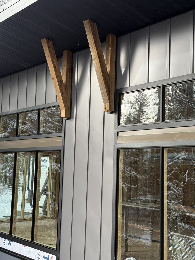 Close-up detail of decorative Y-shaped timber brackets mounted against gray vertical board and batten metal siding above large multi-pane black-framed windows