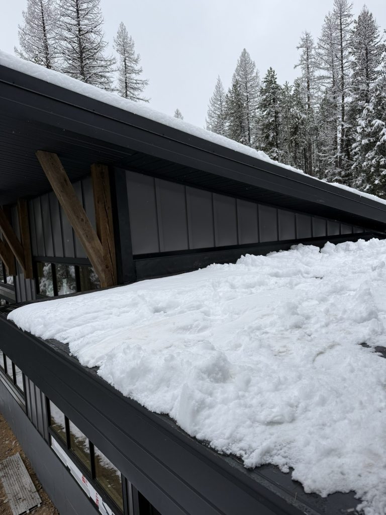 Snow-covered low-slope standing seam metal roof section on modern home with dark metal fascia and timber bracket accent visible at roofline