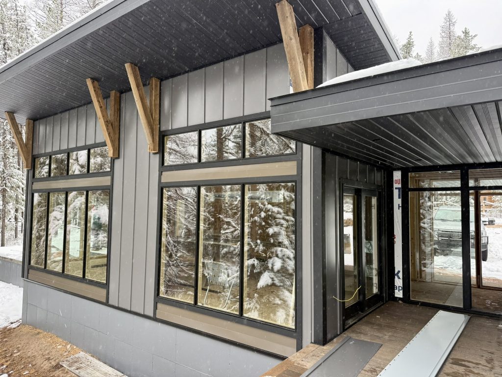 Corner exterior view of modern new construction home with covered entry porch oversized black-framed windows decorative timber brackets and gray board and batten siding in winter