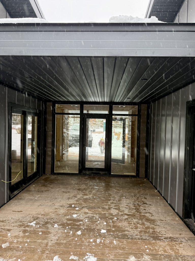 Interior view of covered porch with black metal tongue-and-groove ceiling and full-view glass entry doors opening to a snow-covered deck
