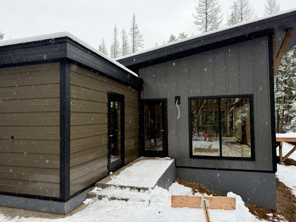 New construction exterior showing gray vertical board and batten siding paired with tan horizontal lap siding accent section and black window and door trim in winter setting