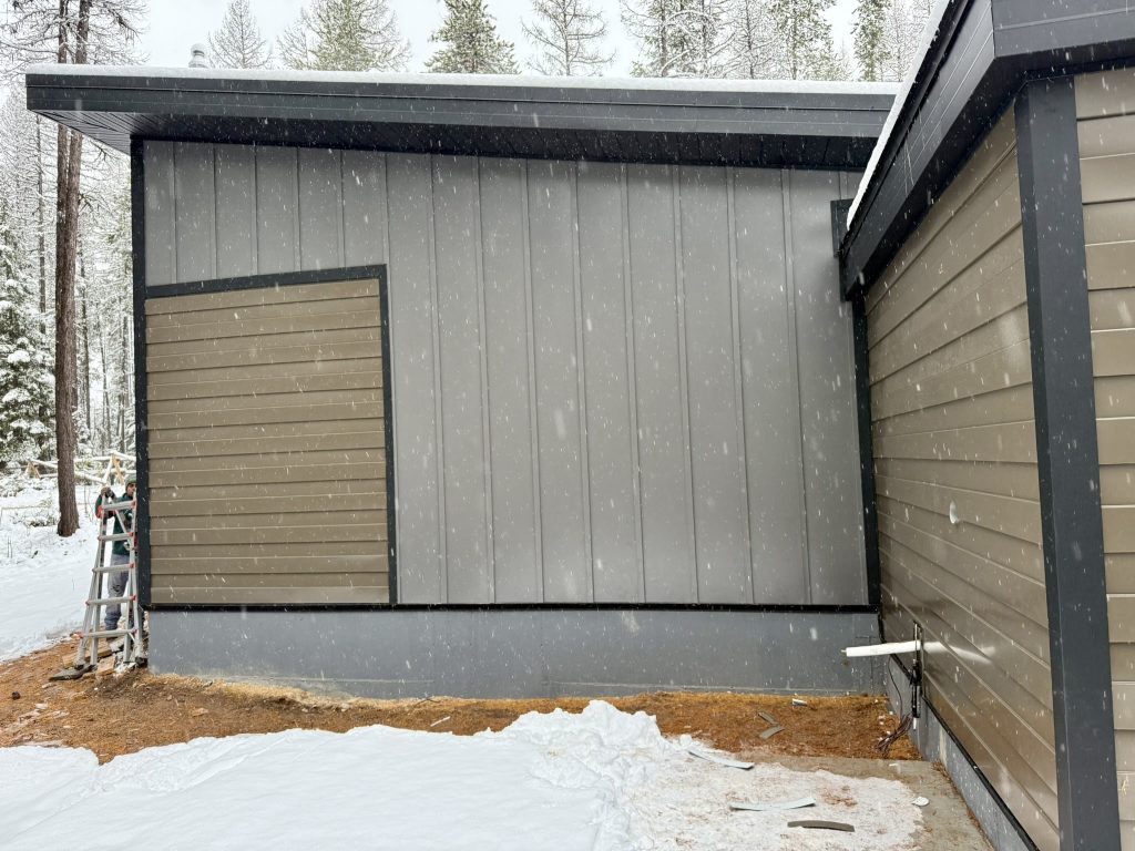 Exterior siding installation in progress on new home featuring gray vertical board and batten panels with tan lap siding accent and black trim in snowy conditions