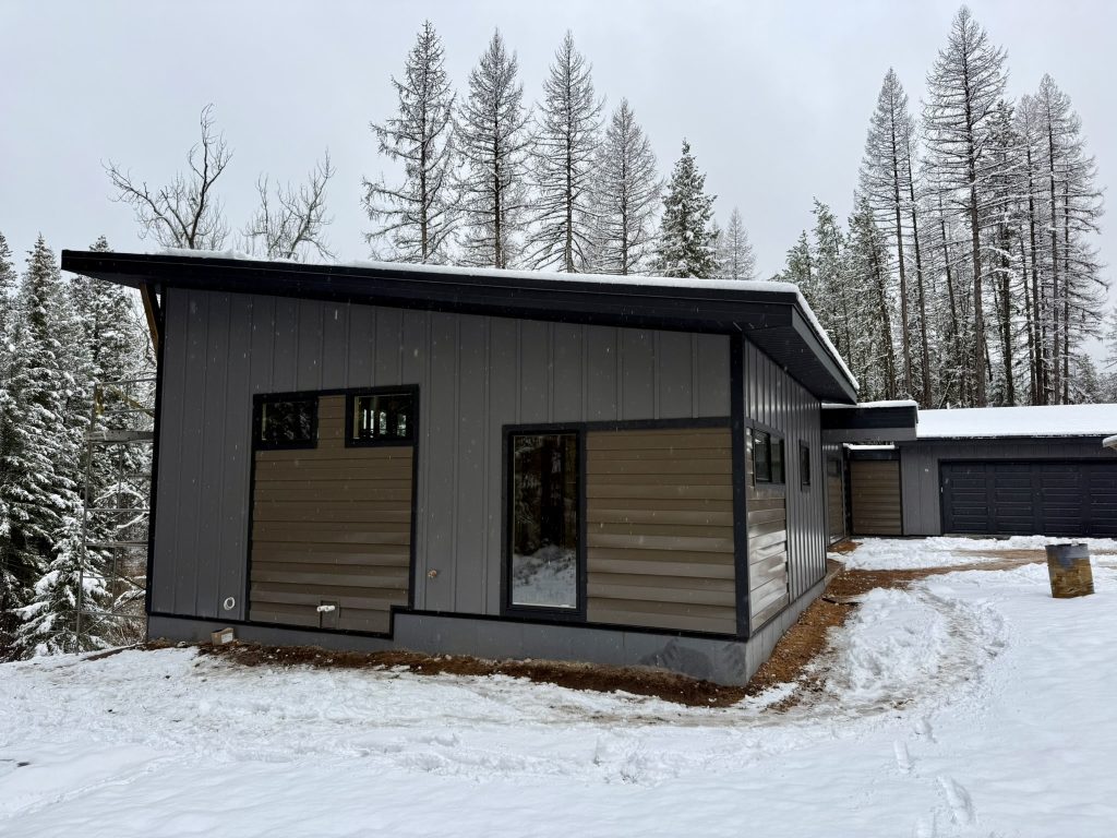 Full exterior view of modern new construction home with single-slope metal roof gray board and batten siding tan horizontal lap accent section and attached garage in winter