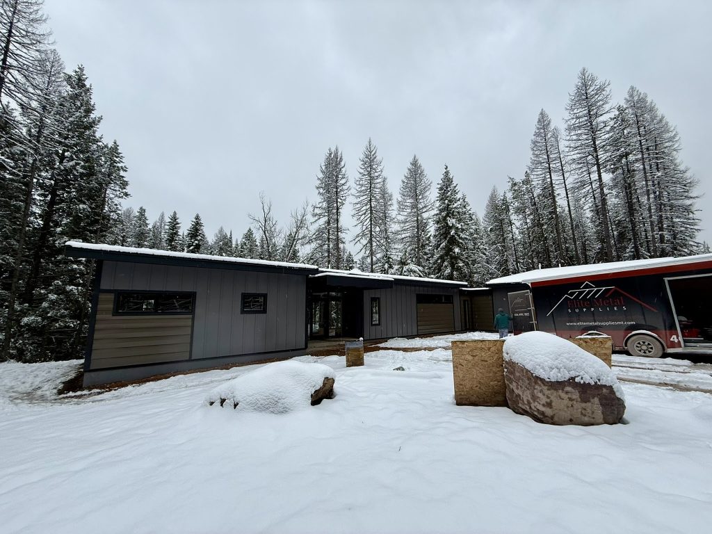 Wide front elevation of modern new construction home with mixed gray and tan siding and low-slope metal roof in snowy forested setting