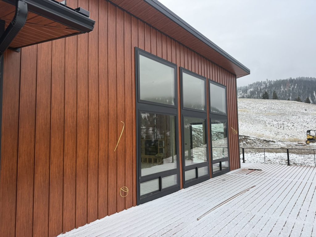 Side exterior of modern home with mahogany red-brown vertical board and batten metal siding large black-framed multi-pane windows and snow-dusted composite deck with cable railing