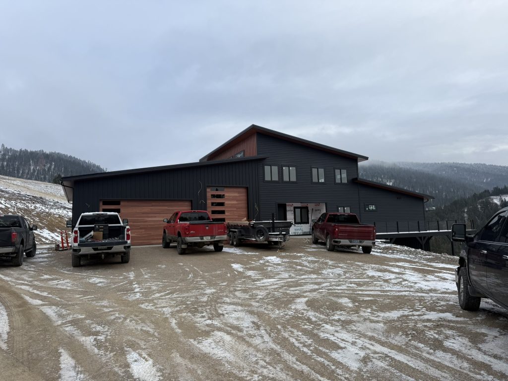 Front view of two-story modern home under construction with dark charcoal lap siding mahogany accent upper gable wood panel garage door and elevated hillside setting in winter
