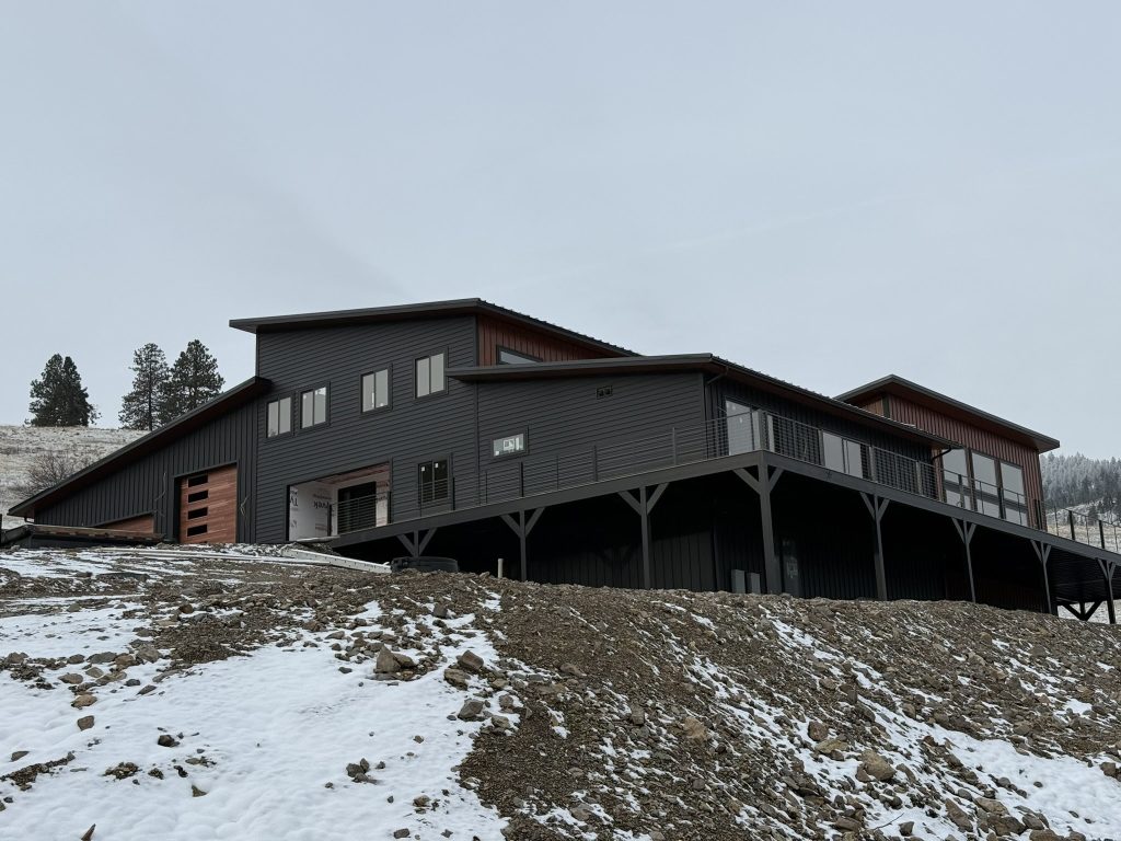 Side view of two-story modern home with dark charcoal siding mahogany upper accent elevated deck with cable railing and forested hillside backdrop in winter