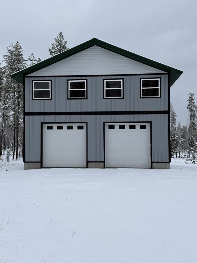 Two-story gray vertical board and batten metal building with dark green trim white garage doors and upper-level windows surrounded by snow-covered forest