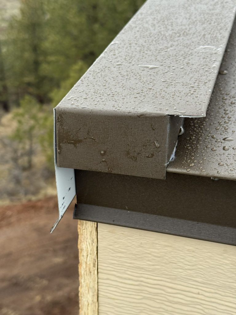 Close-up detail of taupe standing seam metal roof panel edge with water beading at the eave and drip flashing over light wood trim