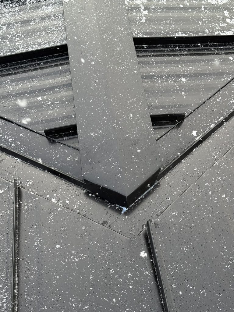 Close-up of complex roof valley and ridge intersection on a black standing seam metal roof with snow speckles showing precision panel and flashing fabrication