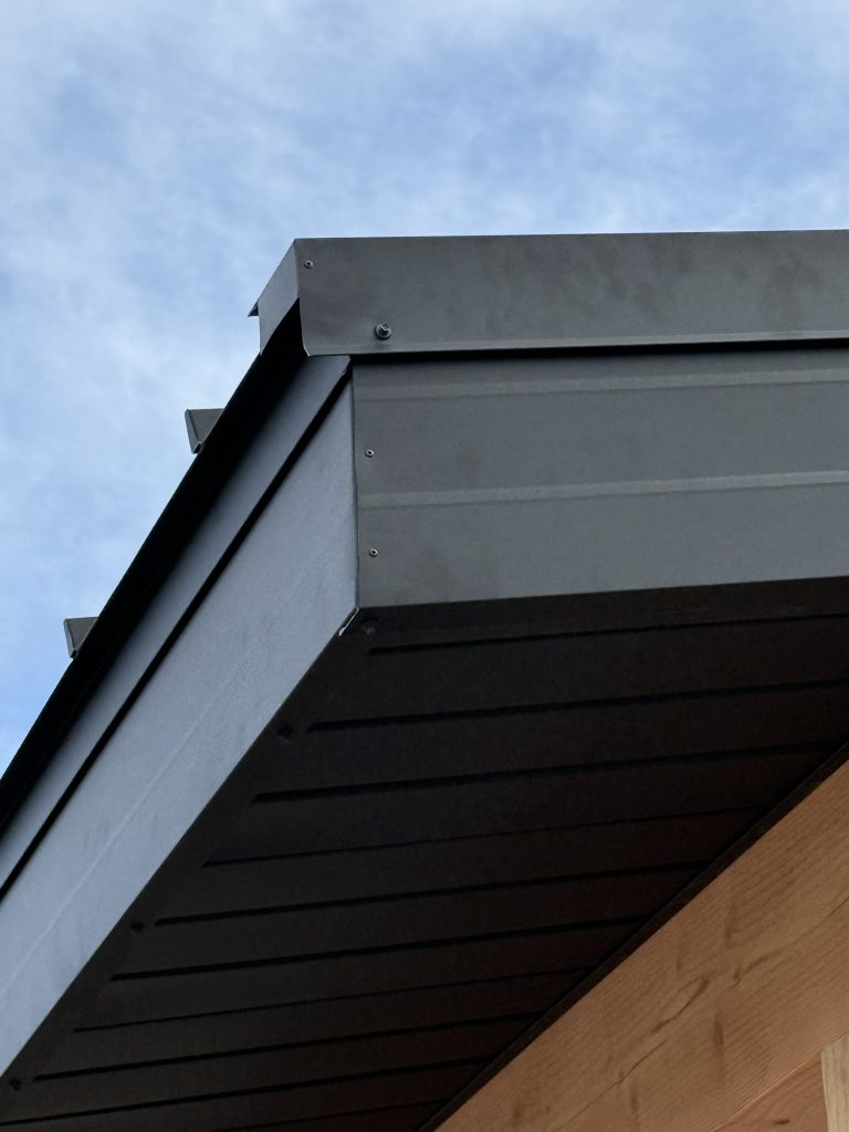 Close-up of dark charcoal metal fascia corner wrap and eave detail against a partly cloudy blue sky showing clean trim fabrication and panel alignment