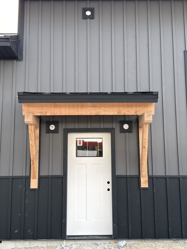 Finished white entry door with natural timber bracket canopy and black metal awning on two-tone gray and charcoal vertical board and batten metal siding with exterior light fixtures