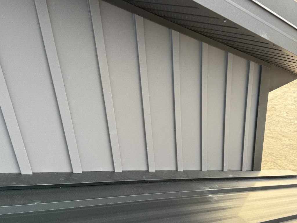 Close-up of light gray vertical board and batten metal siding panels beneath a dark metal eave and soffit showing clean trim installation and panel alignment