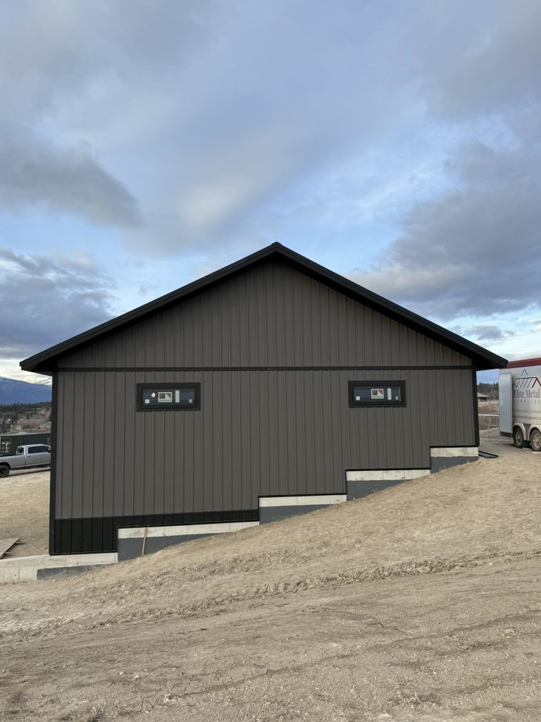 Gable end of dark gray vertical board and batten metal building with black trim detail and mountain range backdrop visible at dusk with Elite Metal Supplies trailer on site