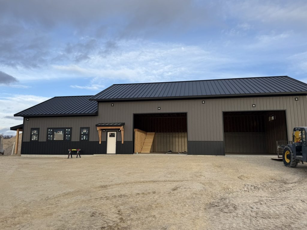 Near-complete front elevation of large barndominium with black standing seam roof two-tone dark and gray board and batten metal siding timber bracket entry door and two commercial bay openings