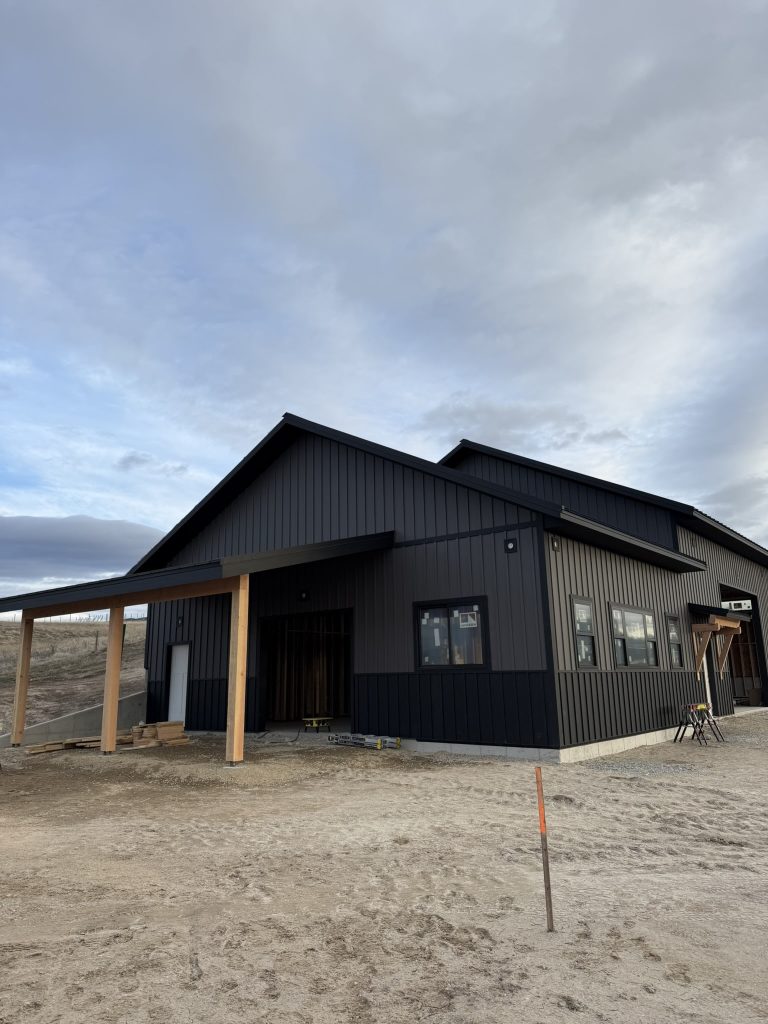 New construction black board and batten metal siding on large barndominium-style building with gabled roof and timber post carport under evening overcast sky