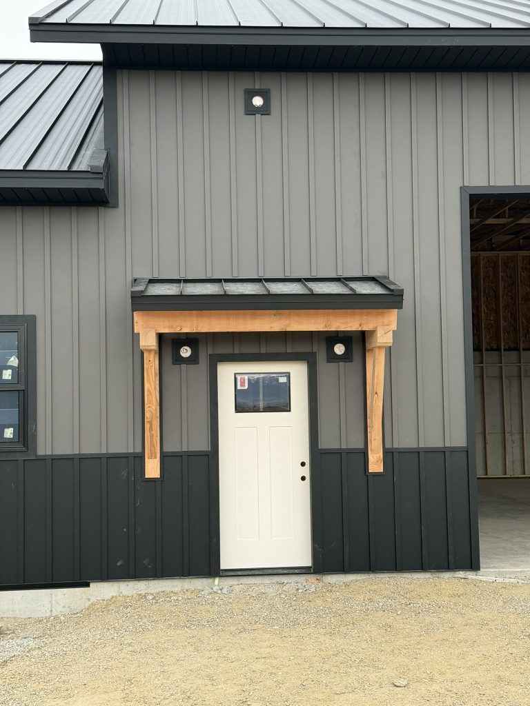 Close-up of white entry door with natural timber bracket canopy and black standing seam metal awning on two-tone gray and charcoal board and batten metal siding