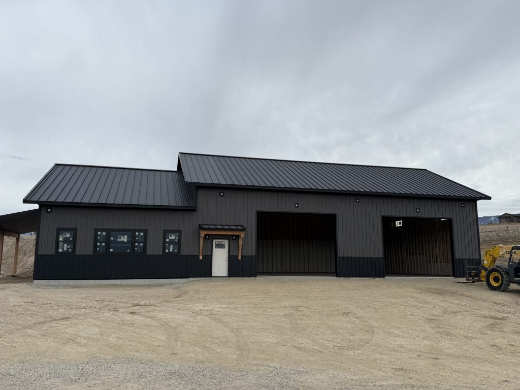 Full front elevation of large barndominium building with black standing seam roof two-tone gray and charcoal board and batten siding timber bracket entry door and two oversized commercial bay doors
