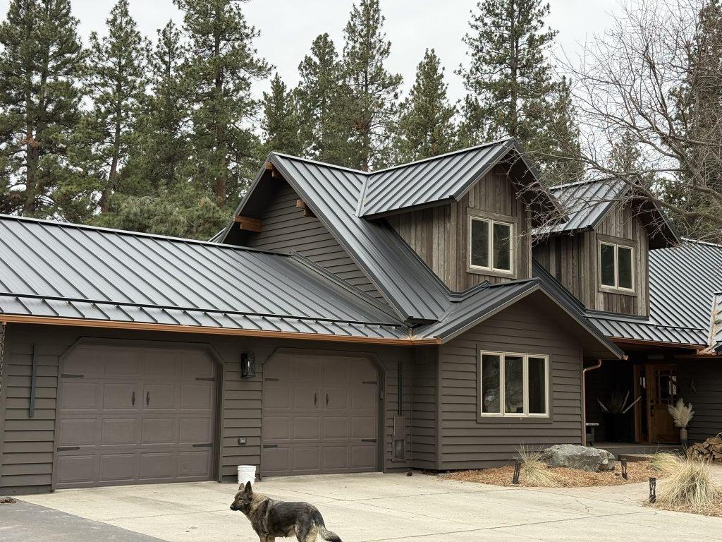 Completed dark charcoal standing seam metal roof on craftsman-style two-story home with multiple gables wood siding and attached two-car garage in forested setting