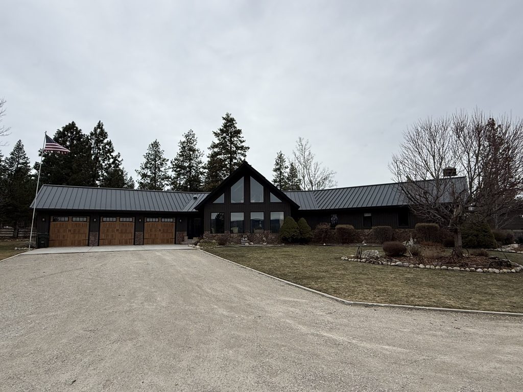Completed dark gray standing seam metal roof on large ranch-style home with wood panel garage doors American flag stone accents and mature pine tree landscaping