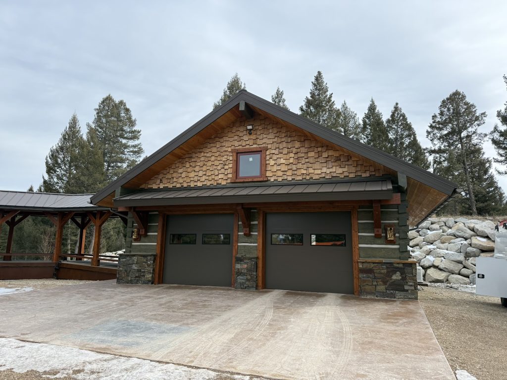 Rustic mountain-style garage with natural cedar shake gable siding stone base dark metal roof standing seam timber frame brackets and two dark gray garage doors